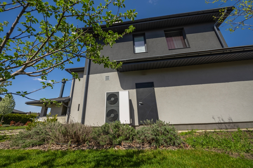 Thermopompe écoénergétique à double ventilateur installée sur le mur extérieur d'une maison contemporaine, entourée d’un gazon frais, d’arbustes et d’arbres en fleurs dans un jardin durable.