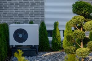 Unité de pompe à chaleur extérieure située dans un jardin bien entretenu d'arbustes et de pierres taillés, placée contre un mur en brique avec un volet de fenêtre fermé, formant une configuration résidentielle économe en énergie. Pompe à chaleur moderne.