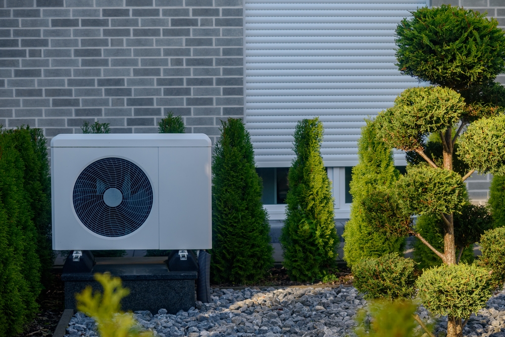 Unité de pompe à chaleur extérieure située dans un jardin bien entretenu d'arbustes et de pierres taillés, placée contre un mur en brique avec un volet de fenêtre fermé, formant une configuration résidentielle économe en énergie. Pompe à chaleur moderne.