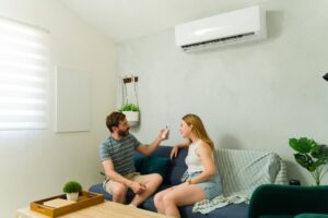 Homme pointant la télécommande du climatiseur sur la femme assise sur le canapé, profitant de l'air frais dans le salon