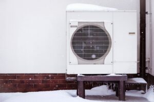 Thermopompe recouverte de neige, installée contre le mur extérieur d’un bâtiment résidentiel en hiver. Système de chauffage moderne et économique pour une maison à faible consommation d’énergie.