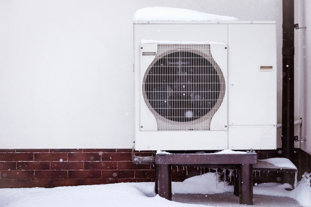 Thermopompe recouverte de neige, installée contre le mur extérieur d’un bâtiment résidentiel en hiver. Système de chauffage moderne et économique pour une maison à faible consommation d’énergie.