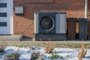 Modern heat pump unit outside brick house in winter. heat Pump unit is installed against the brick wall of a house, next to waste bins and a mailbox.
