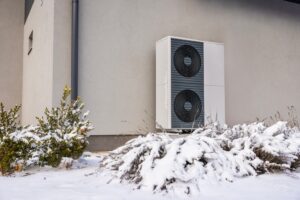 Close-up of a dual-fan outdoor heat pump on a gray wall surrounded by snow-covered bushes, showcasing modern winter energy efficiency. High quality photo