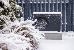 Snow-covered outdoor heat pump unit beside frosted bushes and conifers against a gray metal fence in a winter garden setting