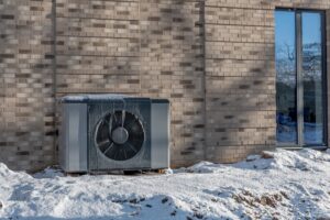 Air source heat pump unit with icicles hanging from casing installed against modern brick building surrounded by snow during winter. Energy-efficient residential heating technology.