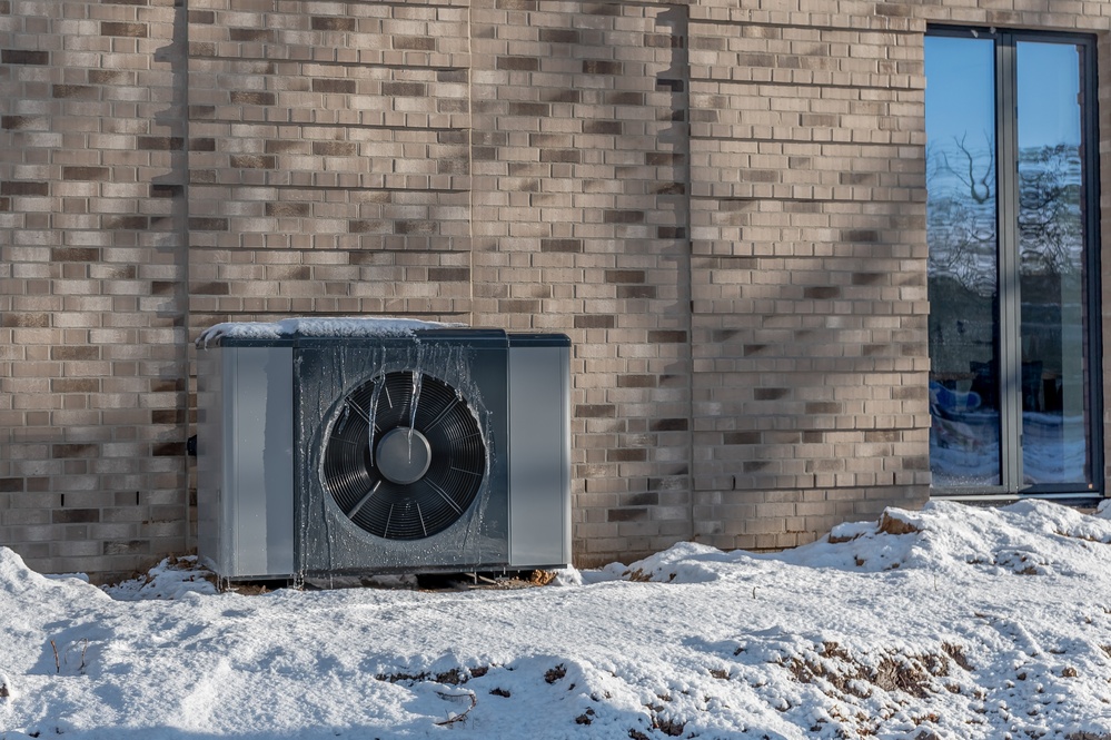Air source heat pump unit with icicles hanging from casing installed against modern brick building surrounded by snow during winter. Energy-efficient residential heating technology.
